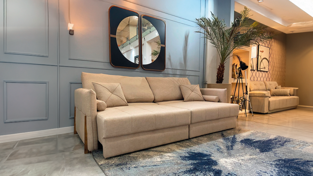 Sofá retrátil bege de dois lugares com almofadas decorativas em uma sala com parede azul acinzentada, boiseries e espelhos decorativos
